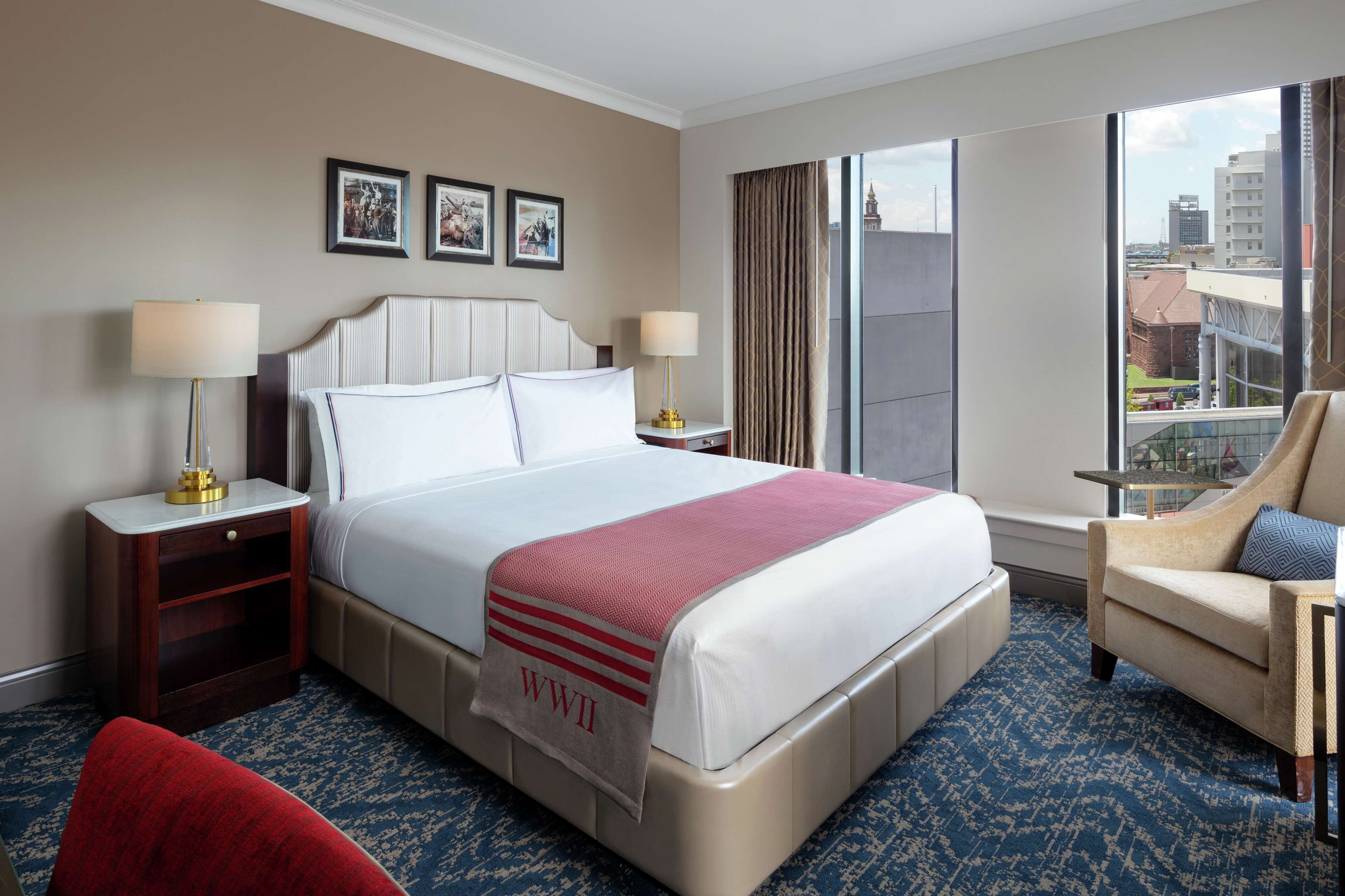 Une chambre d’hôtel avec un grand lit, deux tables de chevet avec des lampes, un fauteuil rouge et une vue sur la ville par une grande fenêtre.