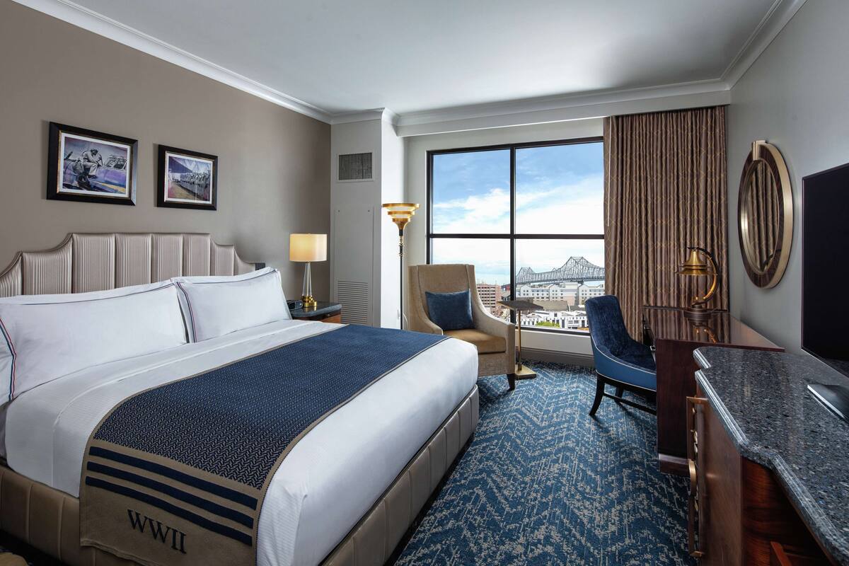 A hotel room with a large bed, a desk, a chair, a lamp, and a view of a cityscape through a large window.