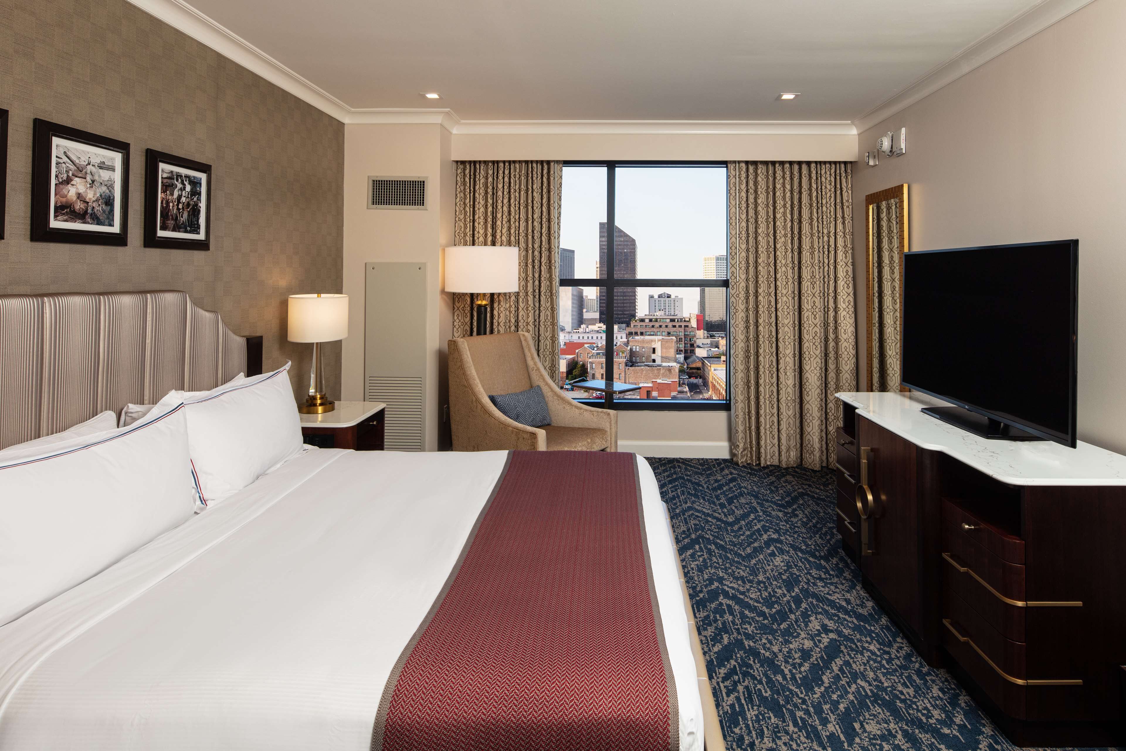 A hotel room with a large bed, a television, a desk, a chair, and a view of the cityscape.