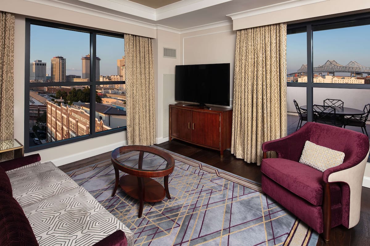 A modern hotel room with a large window offering a city view, a flat-screen TV on a wooden cabinet, a red velvet sofa, and a patterned rug.