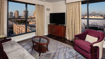 A modern hotel room with a large window offering a city view, a flat-screen TV on a wooden cabinet, a red velvet sofa, and a patterned rug.