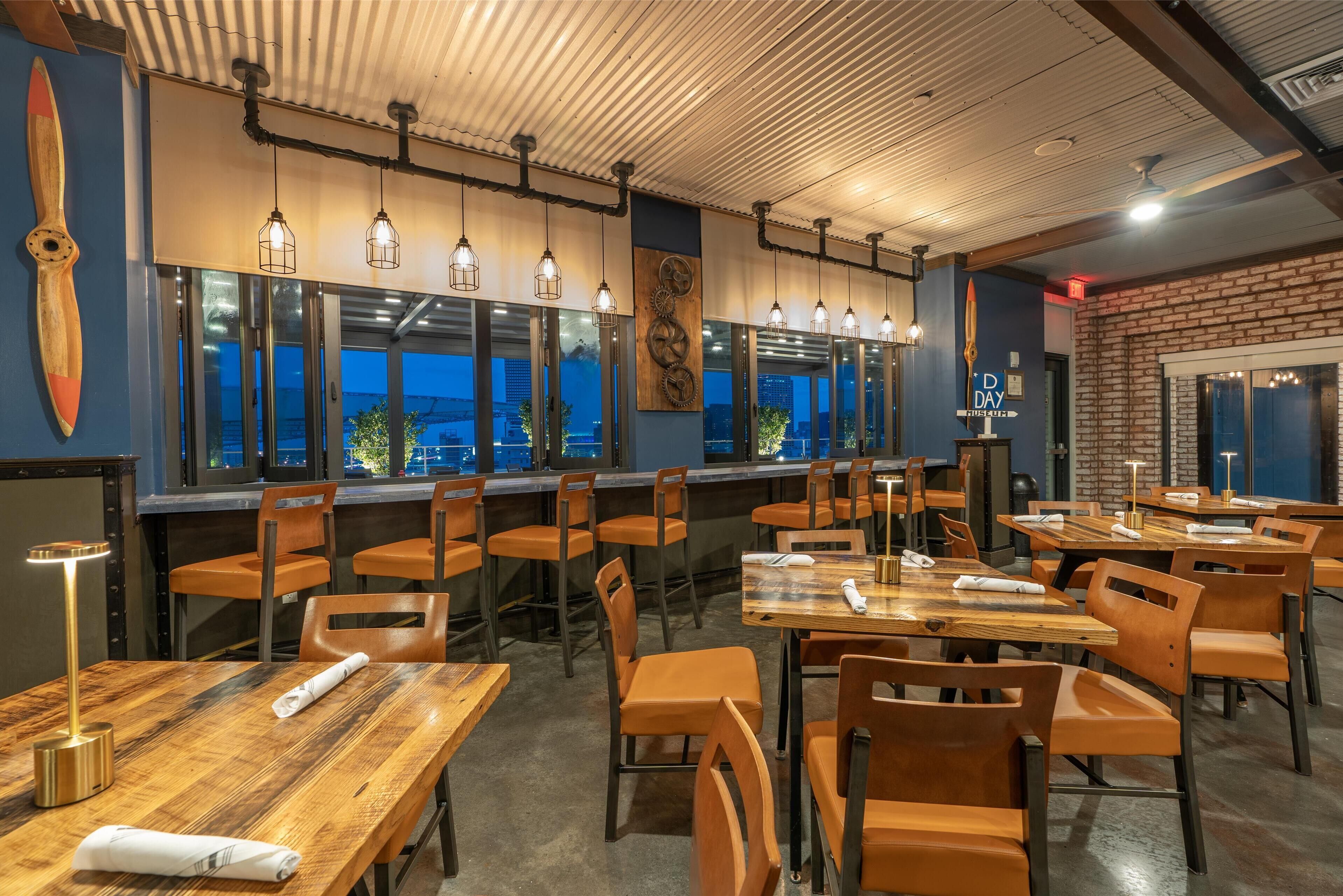 Un restaurante con mesas de madera y sillas naranjas, ventanas amplias con vistas a la ciudad, y un techo con vigas a la vista y luces colgantes.