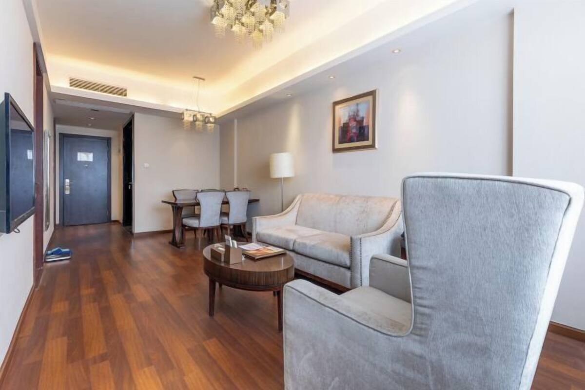 Una sala moderna con un sofá gris, una mesa de centro y un televisor montado en la pared. Se aprecia una zona de comedor con una mesa y sillas al fondo.