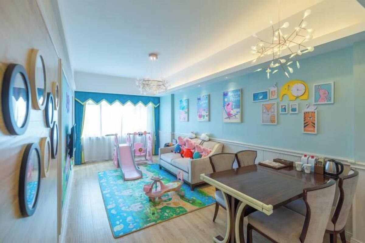 Una habitación con mesa de comedor, sillas, sofá, un rincón de juegos con juguetes y decoración en la pared.