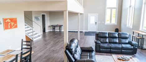 A spacious living area with a high ceiling, a black leather sofa, a wooden coffee table, and a dining table with chairs.