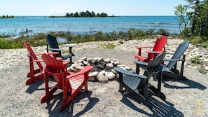 A beach setting with four Adirondack chairs and a small fire pit, surrounded by pebbles and overlooking a calm body of water.
