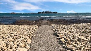 A rocky shoreline with a pebble path leading to the water, a distant tree line, and a clear sky.