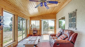 A spacious room with a wooden ceiling, a leather sofa, a desk with a chair, and large windows offering a view of the outdoors. - Sunroom + Sauna! Mtn-View Prescott Valley Home (Prescott Valley)