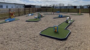 A playground with various play structures, including a windmill and slides, surrounded by a wooden fence and buildings in the background.