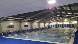 An indoor swimming pool with a blue floor, surrounded by white chairs and tables, under a high ceiling with metal beams and a security camera.