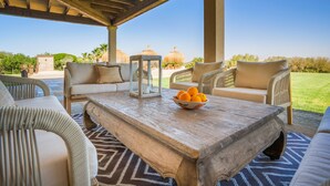 A covered outdoor seating area with wicker furniture, a wooden coffee table with a bowl of oranges, and a clear view of palm trees and buildings in the background. - Great Villa with pool, playground, 5km to the beach Es Trenc, Campos, Mallorca (Campos)