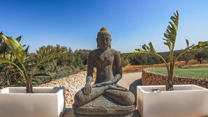 A stone Buddha statue in a meditative pose, surrounded by planters with tropical plants, set against a clear blue sky. - Great Villa with pool, playground, 5km to the beach Es Trenc, Campos, Mallorca (Campos)