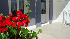 Um edifício branco com persianas azuis, um pátio de concreto e flores vermelhas em primeiro plano.