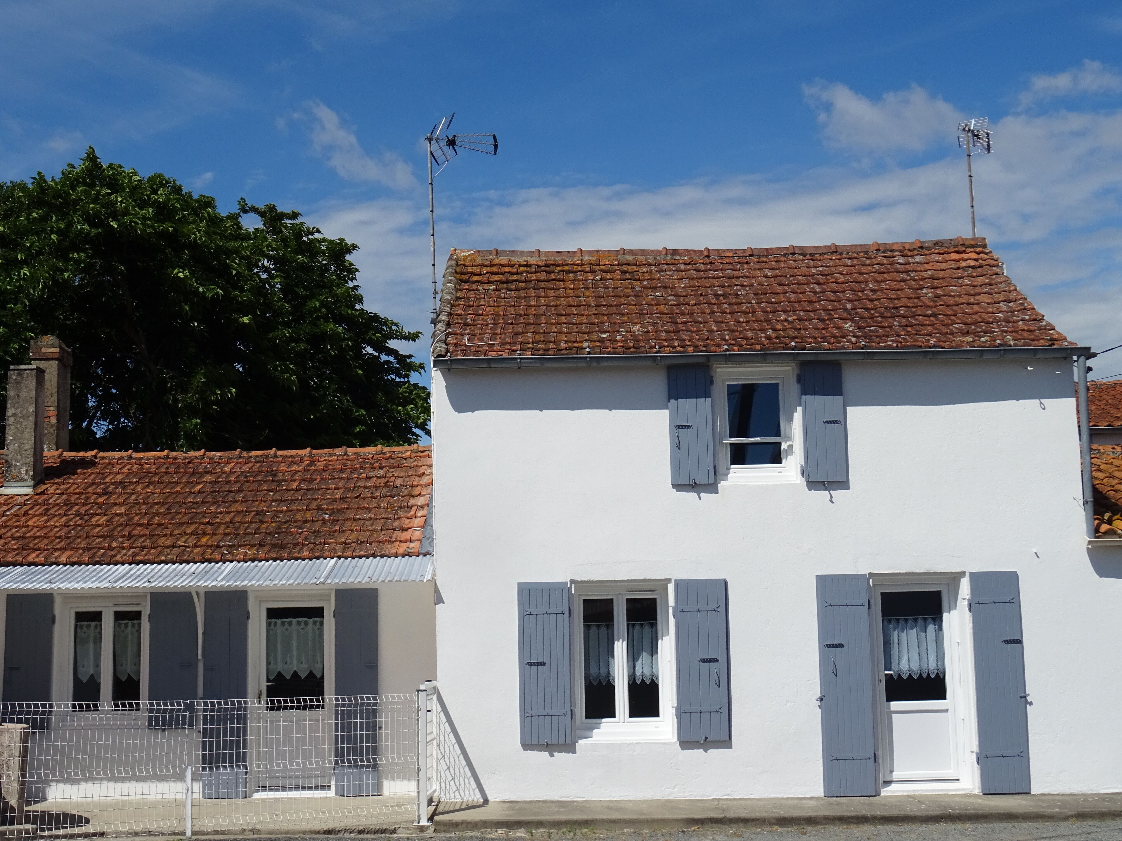 A white building with a tiled roof and shutters, a fenced area, and a tree in the background.
