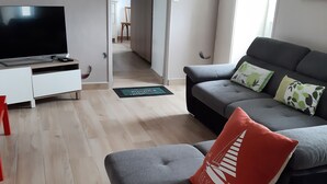 Uma sala de estar moderna com um sofá cinzento, uma almofada vermelha e uma televisão de tela plana sobre um móvel branco.