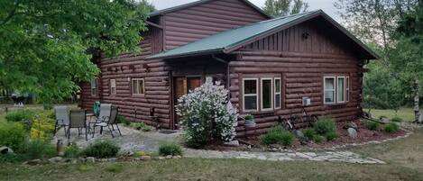 A log cabin with a green roof, surrounded by a garden with a small tree and a patio area with chairs.