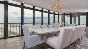 A spacious dining area with a large table and chairs, large windows offering a view of the ocean, and a decorative chandelier. - The Oasis of Orange Beach  1011 (Orange Beach)