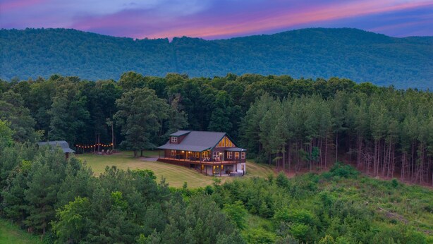 Une grande cabane en rondins dotée d’une véranda qui fait le tour de la maison, entourée d’une forêt dense et de collines.