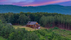 A large log cabin with a wrap-around porch, surrounded by dense forest and hills.