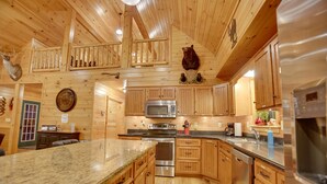 Une cuisine spacieuse dotée de meubles en bois, d’un plan de travail en granit, d’appareils électroménagers en acier inoxydable et d’un plafond en bois avec balcon.