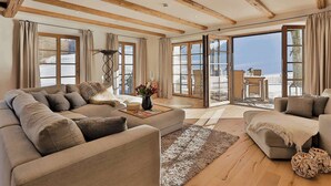 A spacious living room with a large sofa, a smaller armchair, and a coffee table. There are large windows with curtains, a fireplace, and a view of the outdoors. - Chiemsee Chalet (Frasdorf)