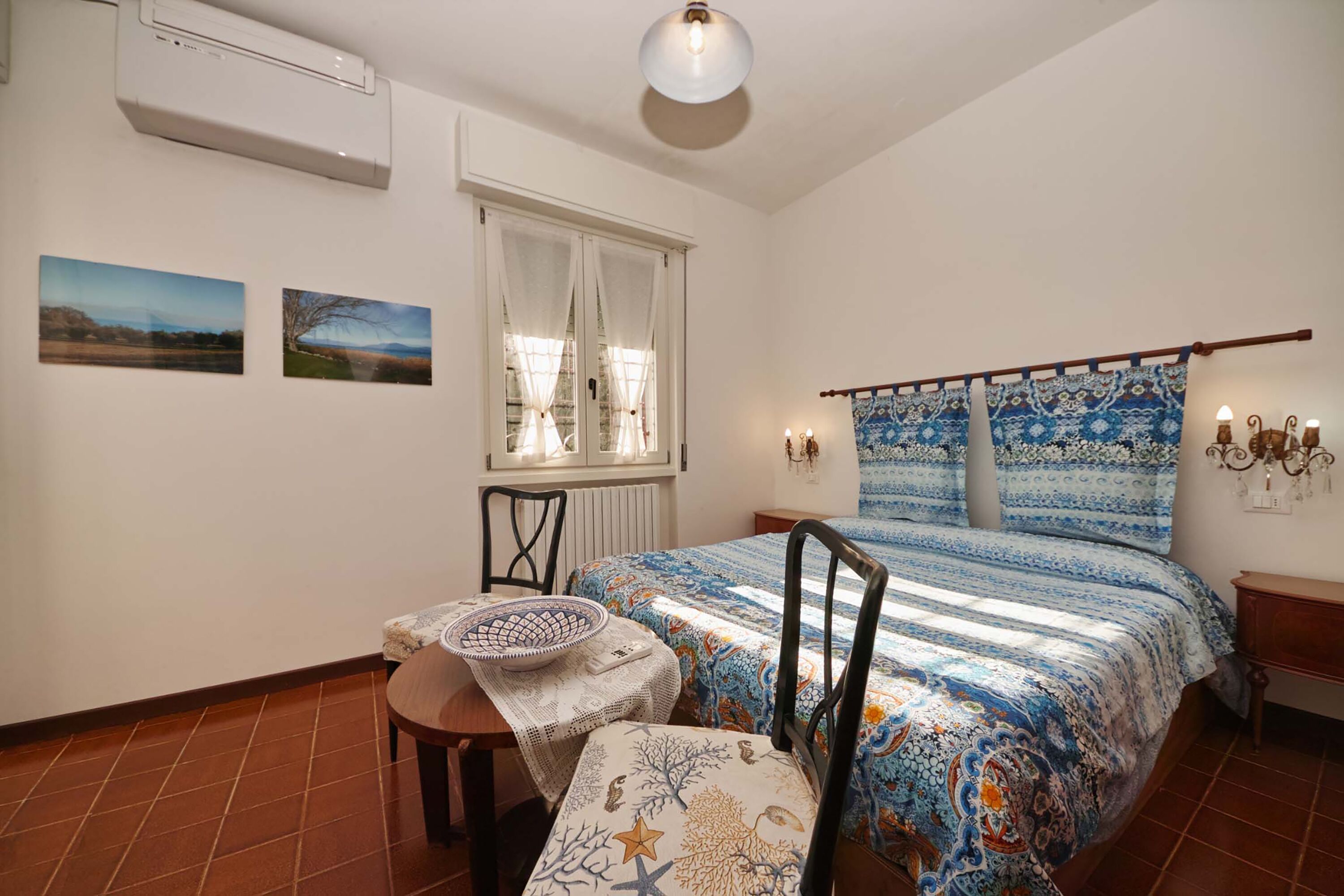 A bedroom with a bed, a table with a chair, a window with curtains, and wall-mounted lights.