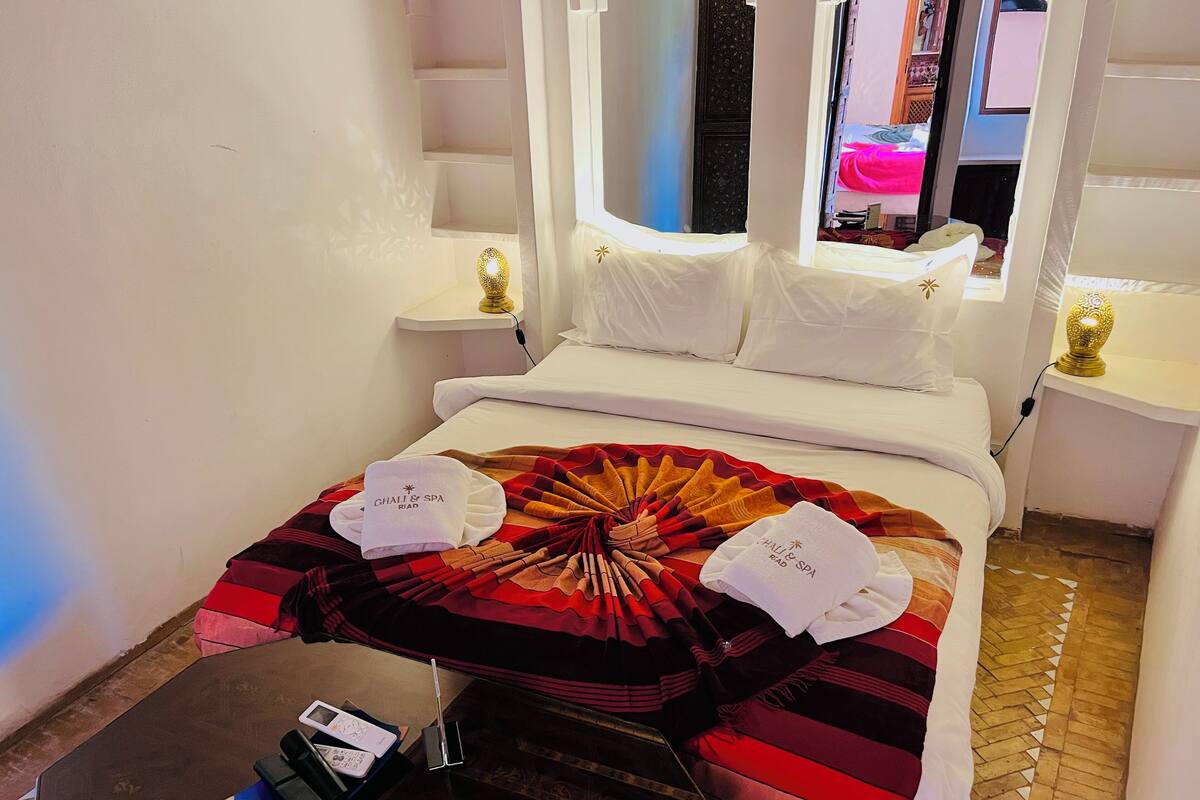 A hotel room with a bed, a bedside table, a lamp, and a view of another room.