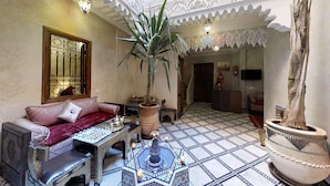 Un interno tradizionale marocchino con pavimento in piastrelle decorate, un divano con cuscini, un tavolino da caffè, un'ampia pianta in vaso e dettagli elaborati al soffitto.