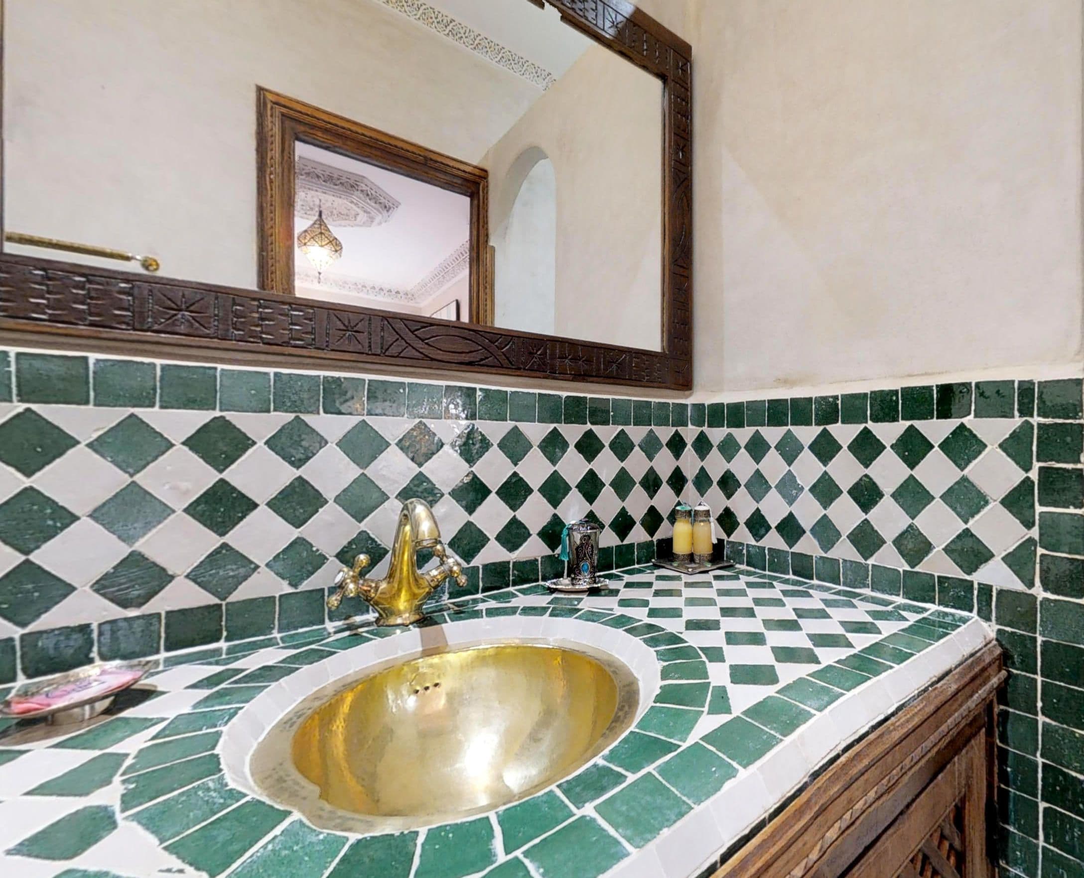 Une salle de bain avec un mur recouvert de carreaux verts et blancs formant un motif à carreaux, un lavabo doré et un meuble en bois.