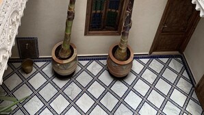 Un cortile con due piante alte in vasi decorati, un pavimento piastrellato con un motivo geometrico e una piccola finestra con cornice scura.