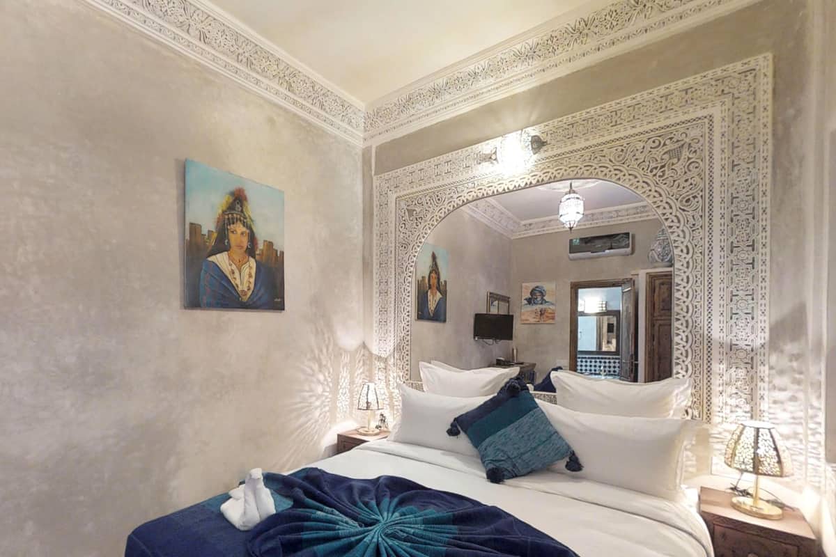 A hotel room with a large bed, a decorative mirror, a painting on the wall, and a nightstand with a lamp.