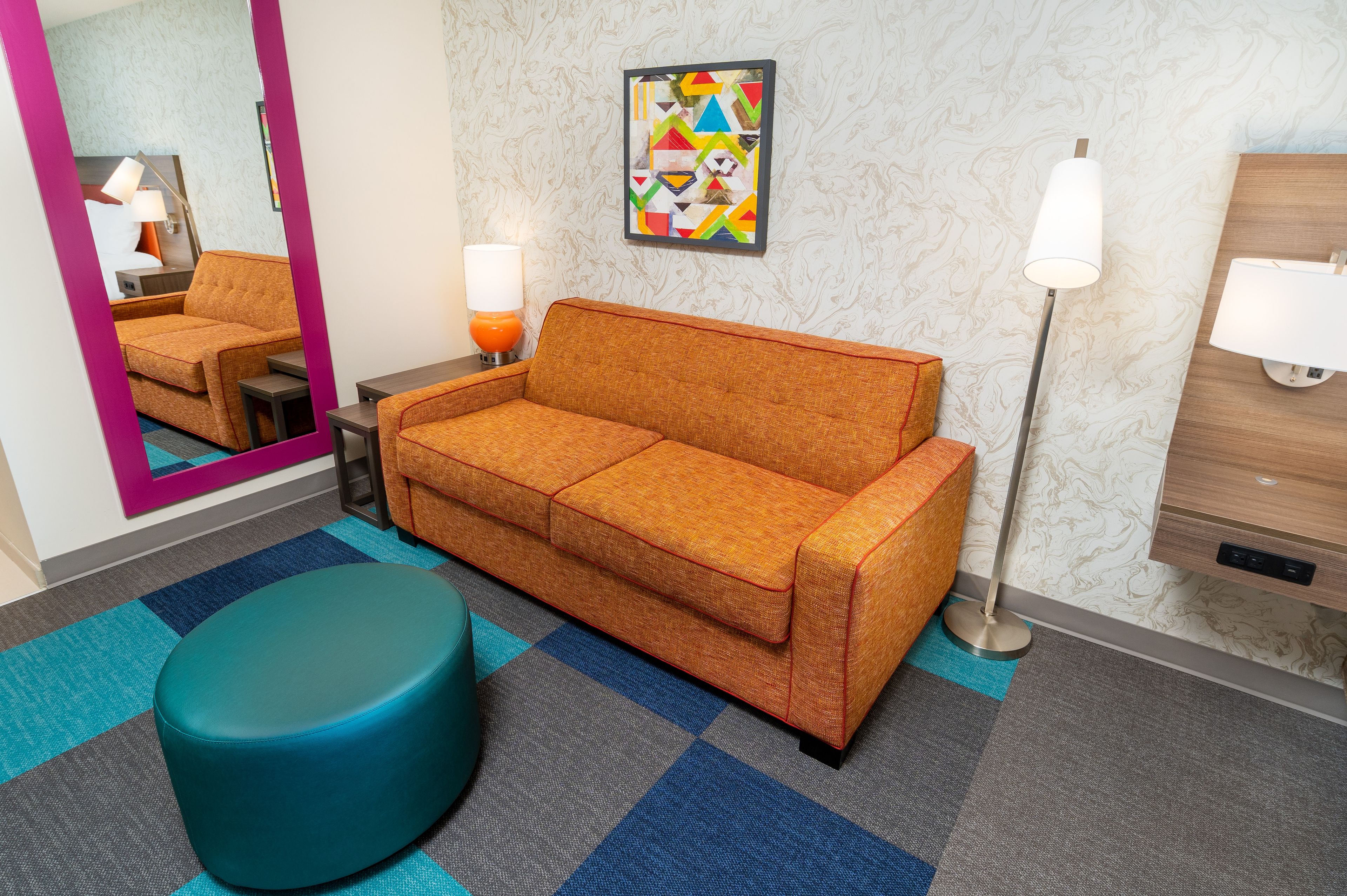 Une chambre d’hôtel moderne avec un canapé, un pouf rond, un miroir, une lampe et une œuvre d’art murale.