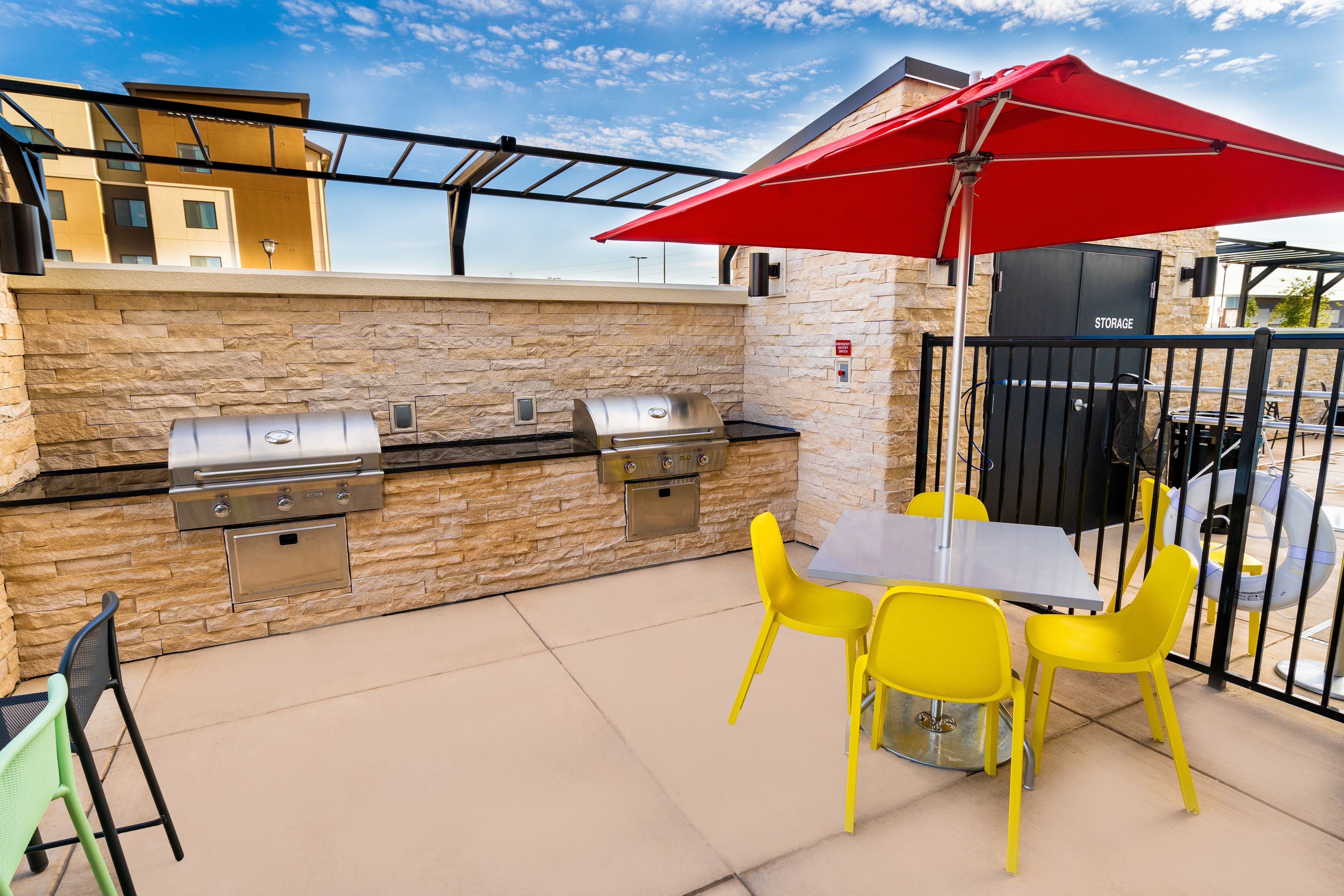 An outdoor patio with a stone wall, a grill, a table, and yellow chairs.