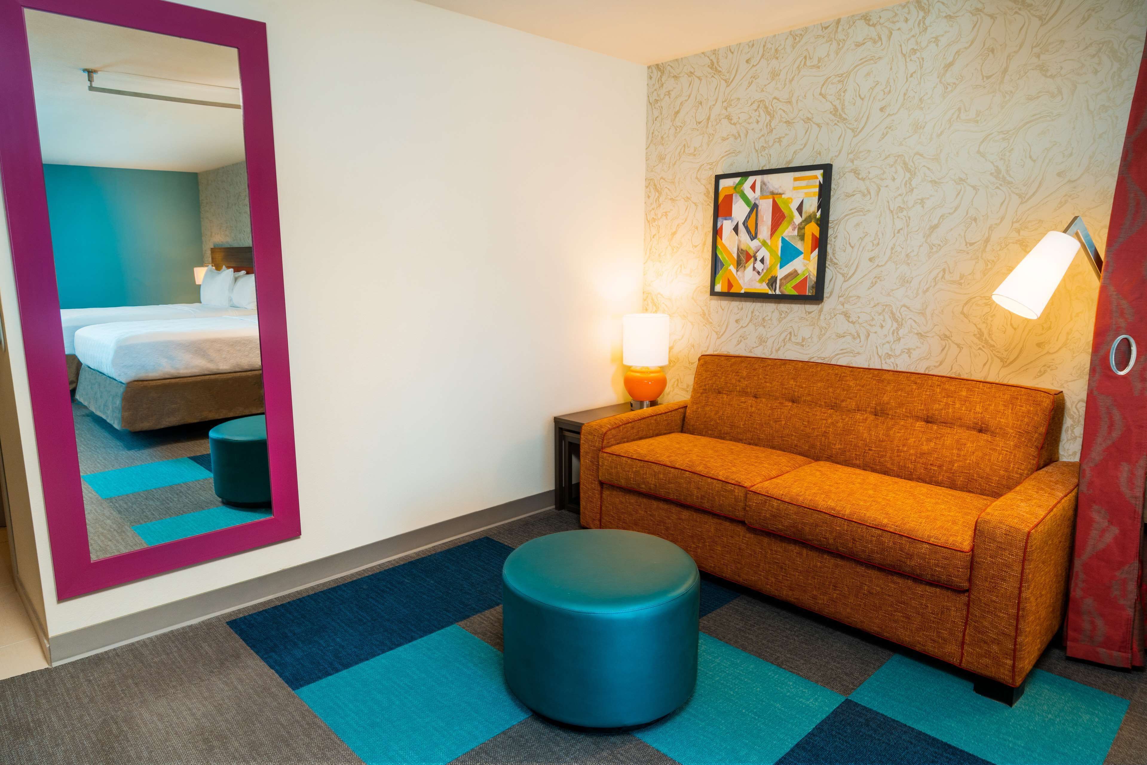 Een hotelkamer met een bed, een sofa, een rond poefje, een spiegel, een wandlamp en een kleurrijk abstract schilderij.