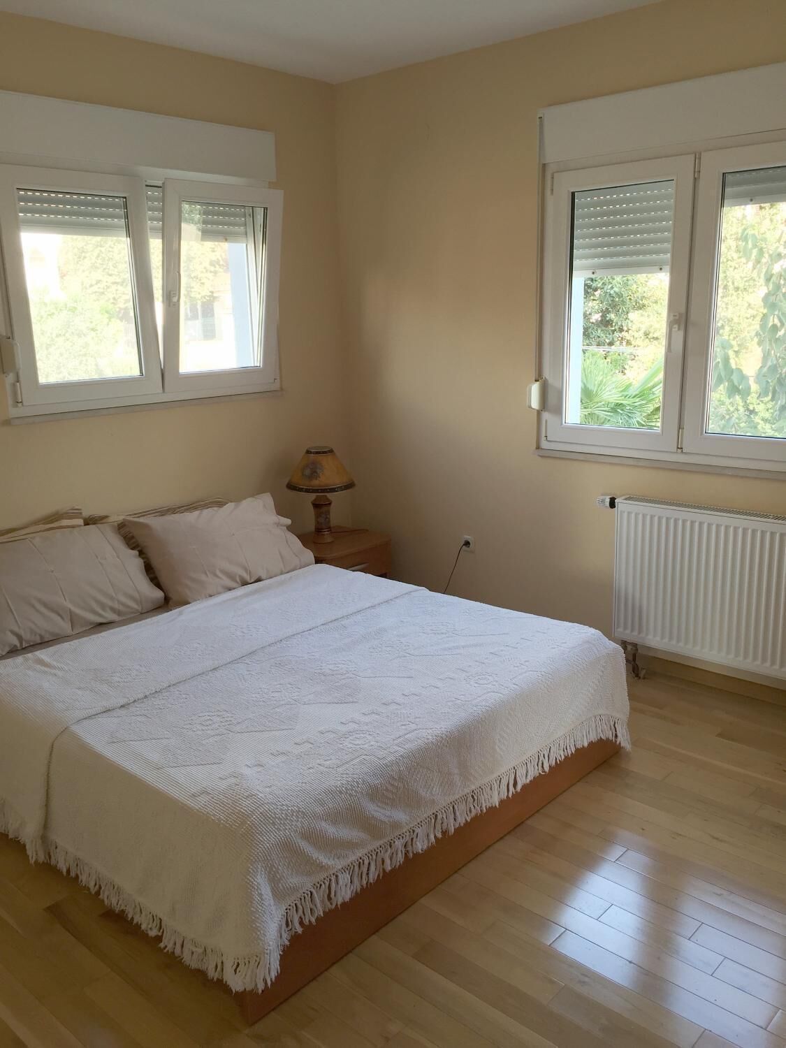 A bedroom with a bed, two windows, a nightstand, and a radiator.