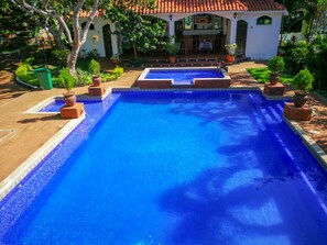 A rectangular pool with a tiled deck surrounding it, a patio area with outdoor seating, and a building with a tiled roof in the background.