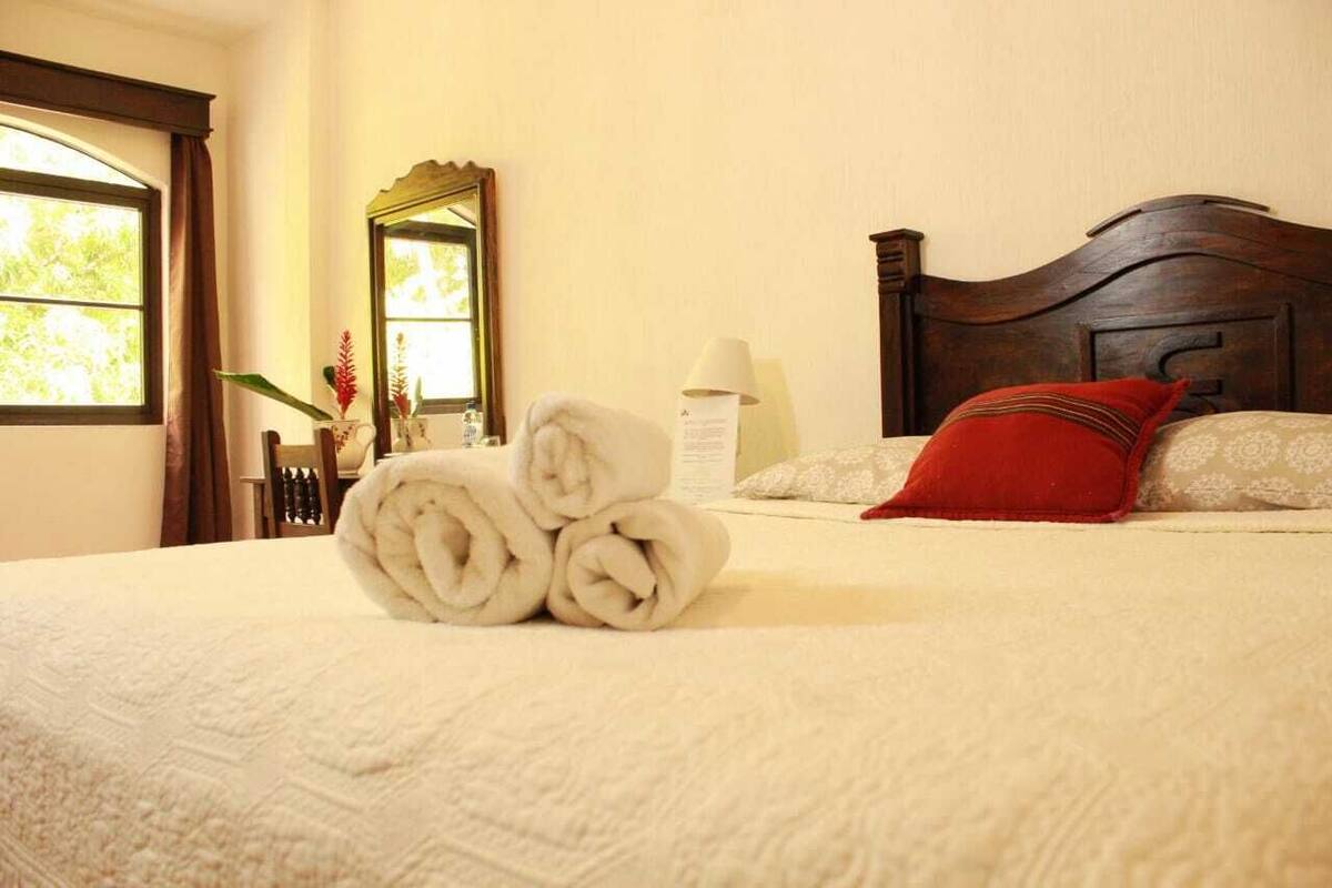 A neatly made bed with rolled towels, a wooden headboard, a mirror, and a window with curtains.