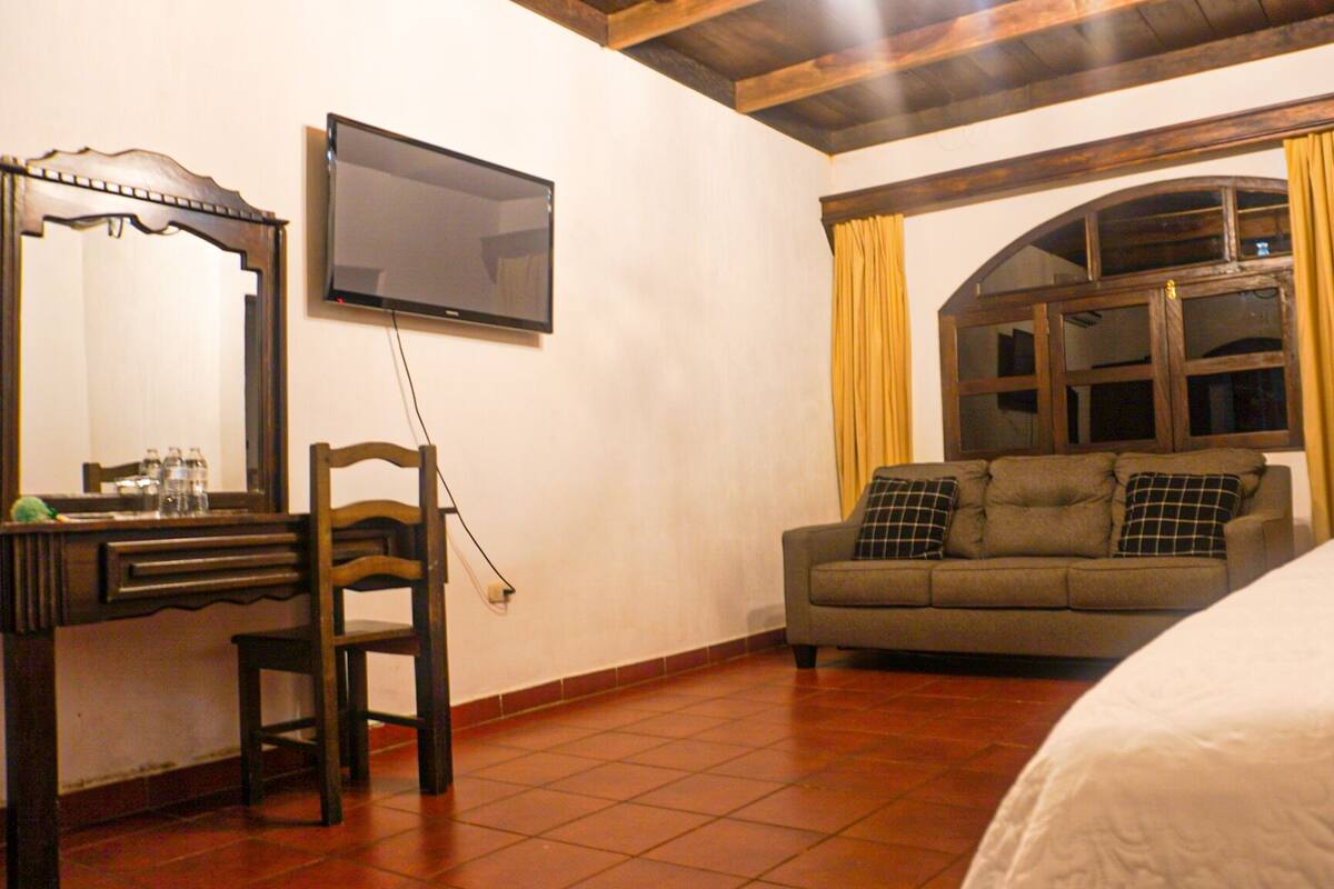 A hotel room with a bed, a sofa, a TV, a desk with a chair, and a window with curtains.