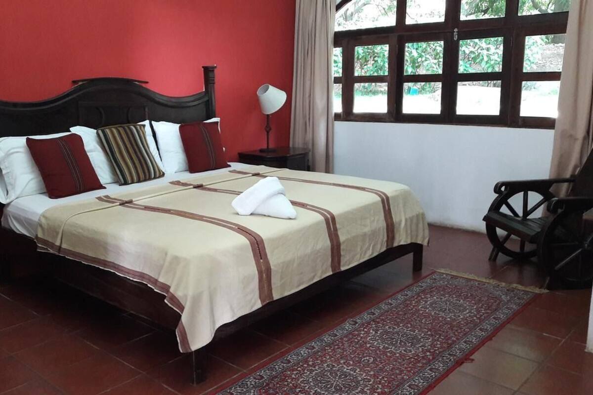 A neatly made bed with a white and beige bedspread, a wooden headboard, and a red wall. There is a window with curtains, a small table with a lamp, and a chair.