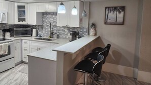 A modern kitchen with white cabinets, a built-in oven, a microwave, and a coffee machine. There is a small island with a sink and bar stools. - Keys stilt cottage lower unit (Naples)