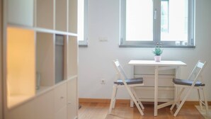 Een kamer met een witte tafel en twee stoelen, een kamerplant op de tafel en een raam met uitzicht op gebouwen.