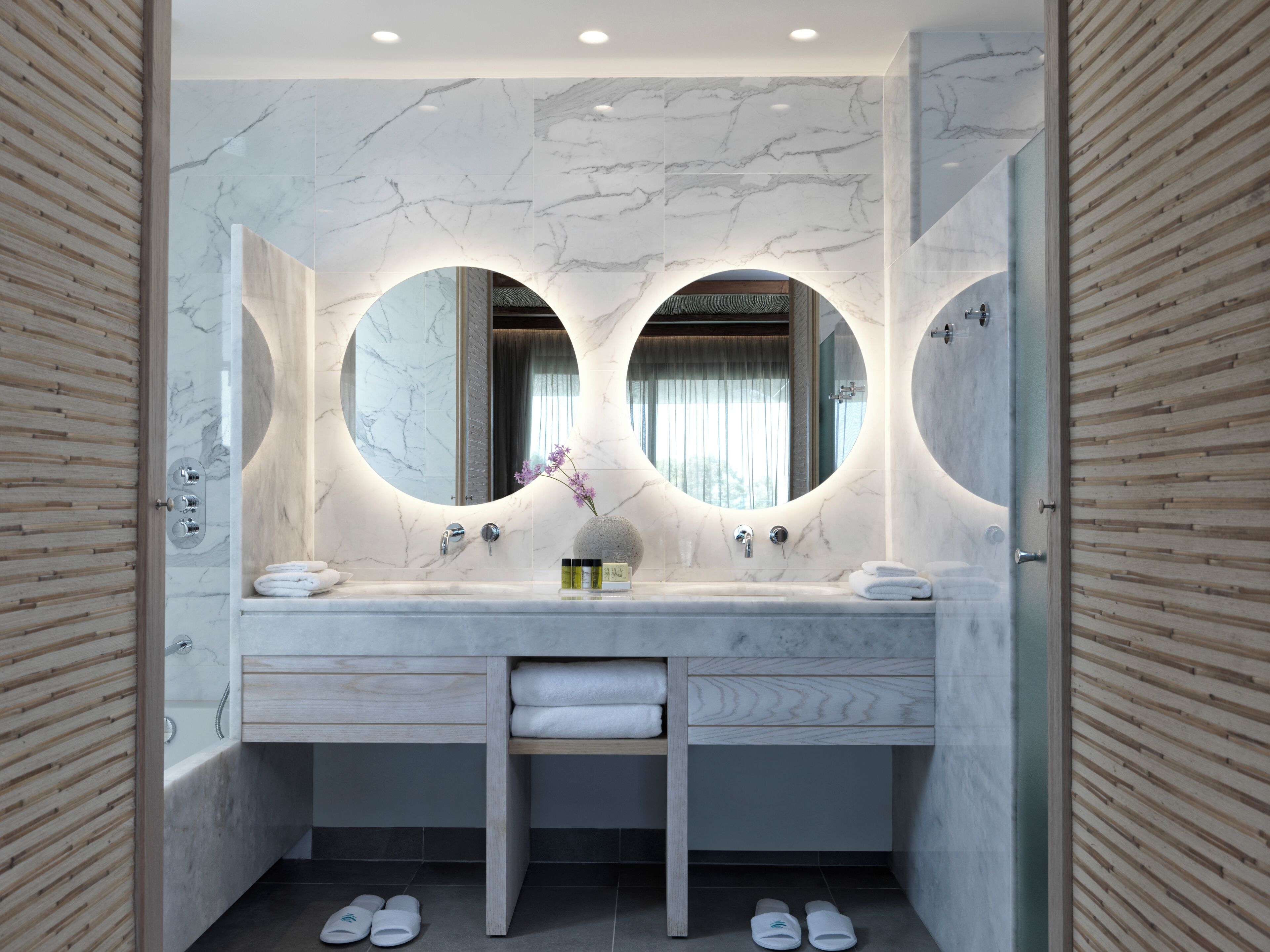 A modern bathroom with a marble vanity, two large mirrors, a wooden bench, and a shower area.