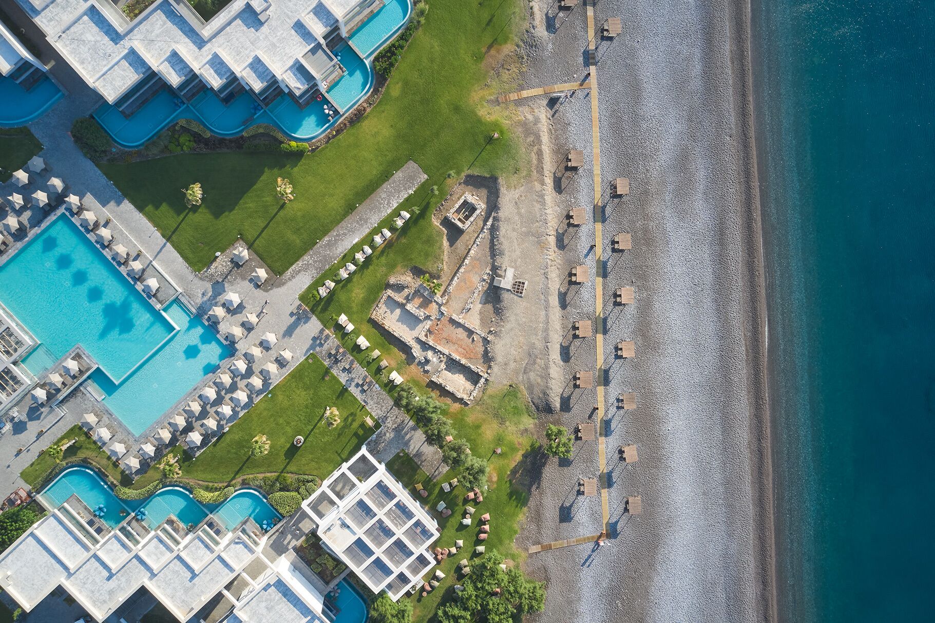 Eine Vogelperspektive zeigt einen luxuriösen Urlaubsort mit mehreren Schwimmbecken, einem Strandbereich und einer felsigen Küstenlinie.