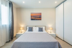 Un dormitorio moderno con una cama grande, dos mesitas de noche, un cuadro con una vista urbana y una panorámica de la ciudad al atardecer.