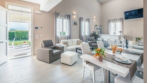 A spacious living area with a dining table set for four, a sofa, and a balcony with a view of greenery.