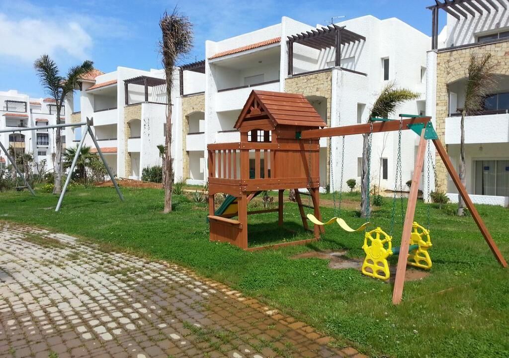 A children's play area with a wooden swing set and a slide, surrounded by a well-maintained lawn and a paved walkway.