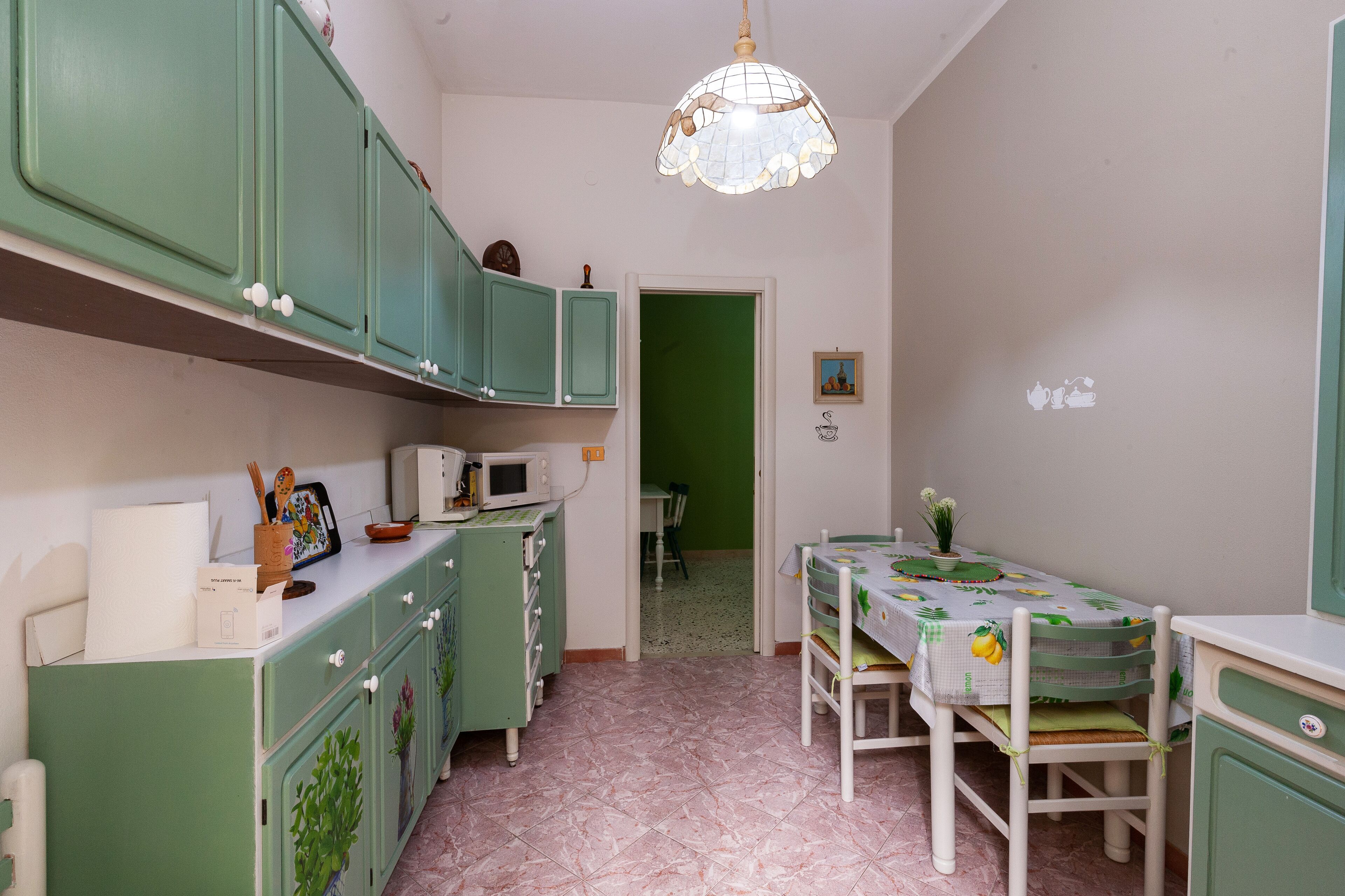 Une cuisine avec des meubles de rangement verts, une table à manger recouverte d’une nappe à motifs de fruits et un luminaire suspendu.