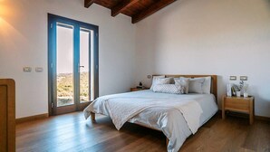 Ein Schlafzimmer mit Holzboden, einem Bett mit weißer Bettwäsche, einem Nachttisch mit Lampe und einem großen Fenster mit Blick ins Freie.