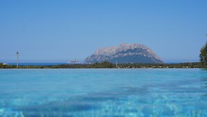 Eine Küstenlandschaft mit einem markanten Felsenberg im Hintergrund, klarem blauen Wasser im Vordergrund und spärlicher Vegetation entlang der Küste.