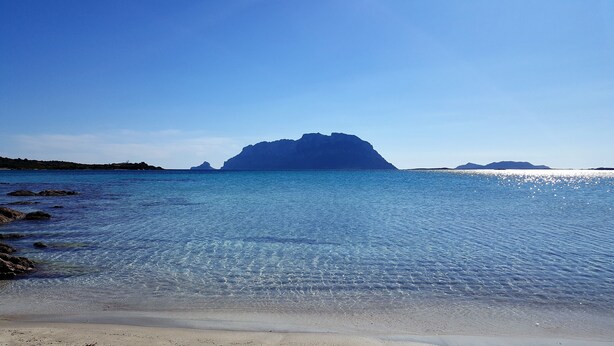 Ein Strand mit klarem, blauem Wasser, einer felsigen Küste und Bergen im Hintergrund.